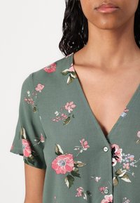 Blusa floreale in tessuto verde con scollo a V, maniche corte e motivi floreali rosa e bianchi. Presenta una chiusura a bottoni sul davanti.