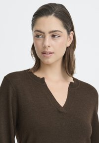 Smeđi pleteni džemper s V-izrezom i rubom sa valovitim dizajnom. Tkanina je teksturirana, s mekanim izgledom i prigodnim krojem.