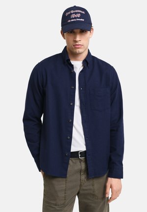 Chemise boutonnière bleu marine avec une poche poitrine, portée sur un t-shirt blanc. Associée à un pantalon vert olive et une casquette bleu marine avec du texte rouge.