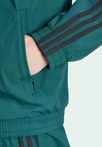 Teal træningsjakke med elastiske manchetter, en lynlåslomme og sorte accentstriber langs ærmerne. Glat, let stoftekstur.