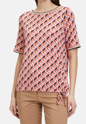 Donna che indossa una maglietta a maniche corte con motivo geometrico multicolore e orlo con coulisse, abbinata a pantaloni marrone chiaro.