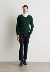 Pulover verde cu decolteu în V, fabricat dintr-un material moale, asociat cu o cămașă albă cu guler și pantaloni negri, purtat cu pantofi negri. Design simplu și modern.