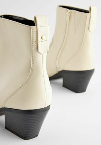 Beige anklar stövlar i syntetläder med en strukturerad svart klack. Har en sidodragkedja och ett litet dekorativt band med en guldklick.