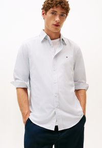 Camicia a maniche corte azzurra a fantasia con maniche arrotolate, colletto a bottoni e un piccolo logo sul petto. Indossata sopra una maglietta bianca.