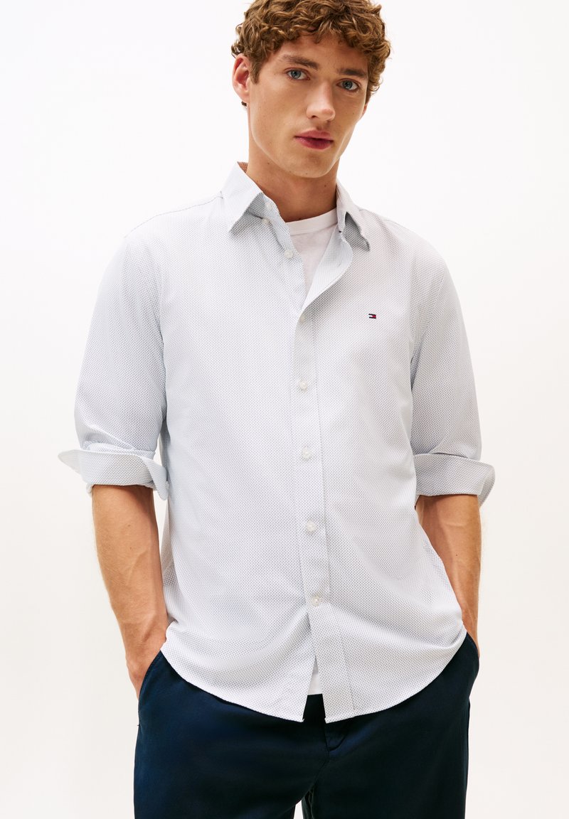 Lichtblauw shirt met een patroon, korte opgerolde mouwen, een knoopsluiting kraag en een klein logo op de borst. Draagt over een wit T-shirt.