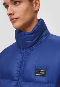 Blauwe pufferjack met een hoge kraag, ritssluiting aan de voorkant en gestructureerde stof. Voorzien van een label op de linkerkant van de borst met coördinaten en tekst.
