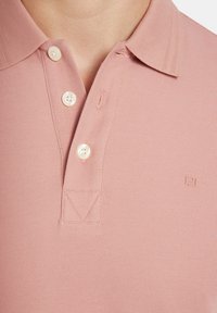 Koszula polo z miękkiego różowego materiału o fakturowanym wykończeniu dzianiny. Posiada zapięcie na guziki, trzy guziki i małe logo w prawym dolnym rogu.