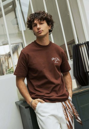 Jeune homme aux cheveux bouclés portant un t-shirt graphique marron et un pantalon blanc avec une écharpe marron à motifs nouée à la taille, debout à l’intérieur.