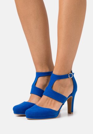 Chaussures à talons hauts en suede bleu avec une plateforme. Présentent des côtés découpés et une bride de cheville avec une boucle en argent. Texture lisse, design épuré.