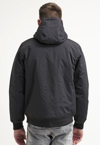 Giacca nera con cappuccio, caratterizzata da una finitura liscia e opaca, polsini e orlo elasticizzati. Presenta un design semplice senza motivi visibili.