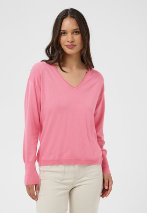 Femme aux longs cheveux bruns portant un pull rose à col en V et un pantalon beige clair, debout devant un fond blanc uni.