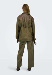 Giacca verde oliva con pannelli in pelle sulle spalle e frange sul retro; abbinata a pantaloni verde oliva coordinati. Tessuto morbido e vestibilità rilassata.