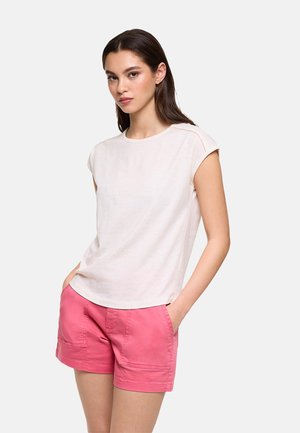 Mujer vestida con una camiseta blanca sin mangas y pantalones cortos rosas, de pie con las manos en los bolsillos contra un fondo claro y liso.