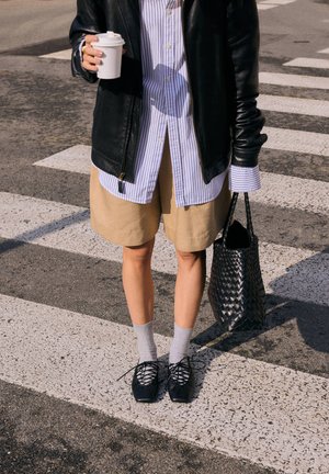 Personne debout sur un passage piéton portant une veste en cuir, une chemise rayée, un short beige, des chaussettes grises, des chaussures noires, tenant une tasse de café et un sac noir tressé.