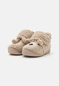 Chaussons en beige tout doux avec une surface texturée, présentant une forme arrondie, des détails de visage d'animal et une semelle en caoutchouc flexible.