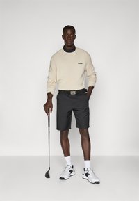 Helle beige Sweatshirt mit "BOSS"-Logo, schwarze Shorts und weiße Sneakers. Hält einen Golfschläger. Glattes Material und sportliche Passform.