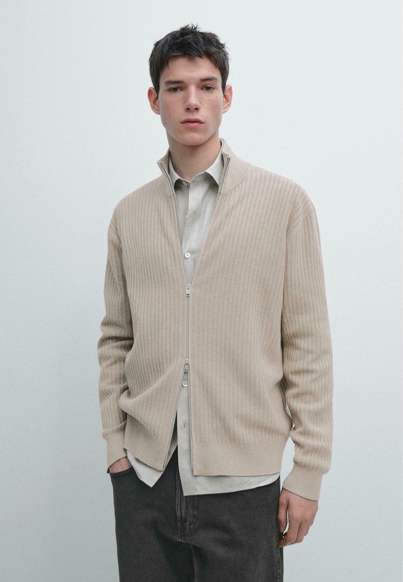Beżowy prążkowany kardigan z wysokim kołnierzem i srebrnym zamkiem błyskawicznym. Nosi się go na jasnoszarą koszulę na guziki i ciemnoszare spodnie.