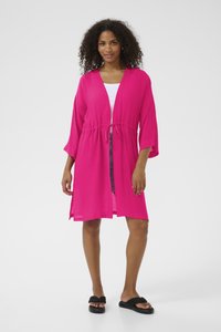Cubrepechos ligero de estilo kimono en color rosa, con cintura ajustable, mangas 3/4 y aberturas laterales, llevado sobre una blusa blanca y combinado con sandalias negras.