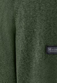 Zelený pletený svetr s texturovaným povrchem. Obsahuje tkaninu s vazbou, žebrované detaily a černou nášivku s bílým textem.