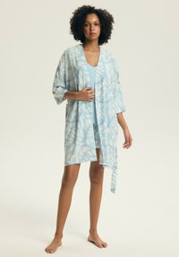 Žena stojaca bosá, oblečená v svetlomodrých šatách a zodpovedajúcom kvetinovom župane s voľnými rukávmi a opaskom v páse.