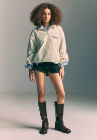 Lysebeige fleecepullover med halvlange lynlås og sort tekst; stylet med denimshorts og brune knælange støvler med spældetalje.