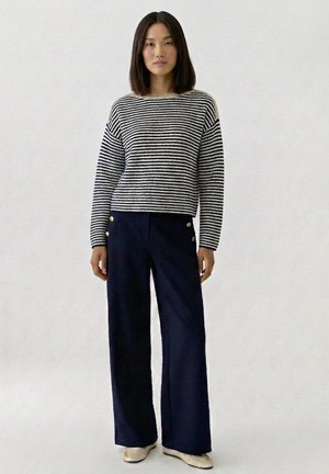 Vrouw staand, gekleed in een zwart-wit gestreepte trui, marineblauwe wijde broek met gouden knopen en beige platte schoenen op een lichte achtergrond.