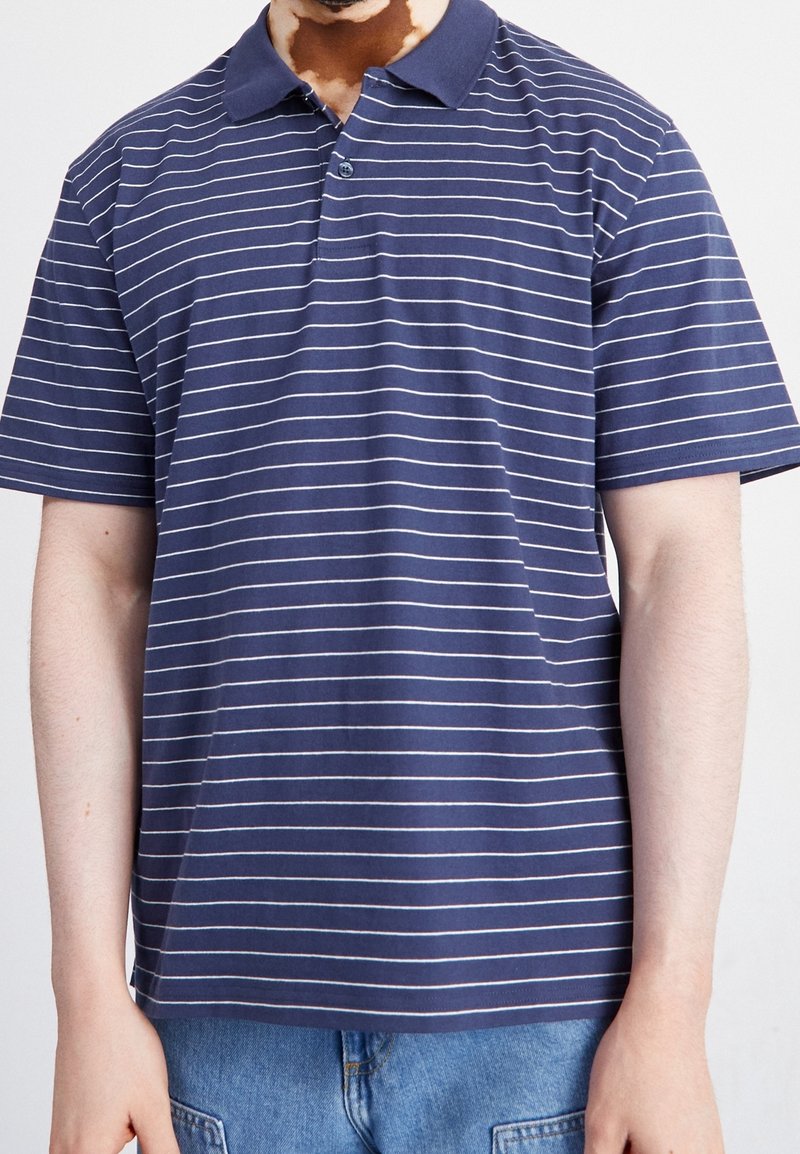 Homme portant un polo bleu marine à manches courtes avec de fines rayures horizontales blanches et un jean bleu clair, debout devant un fond clair.
