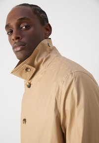 Beige lichtgewicht trenchcoat met een hoge kraag, dubbel-knopsluiting en een zachte textuur. Versierd met glanzende hardware-details.