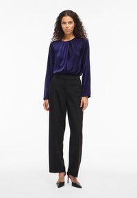 Kvinna som står i svarta vida byxor, en marinblå långärmad blus och svarta spetsiga högklackade skor mot vit bakgrund.