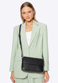 Zwarte textuur schoudertas met reliëfpatronen, verstelbare band en klepsluiting. Draagt een lichtgroene blazer en witte top.