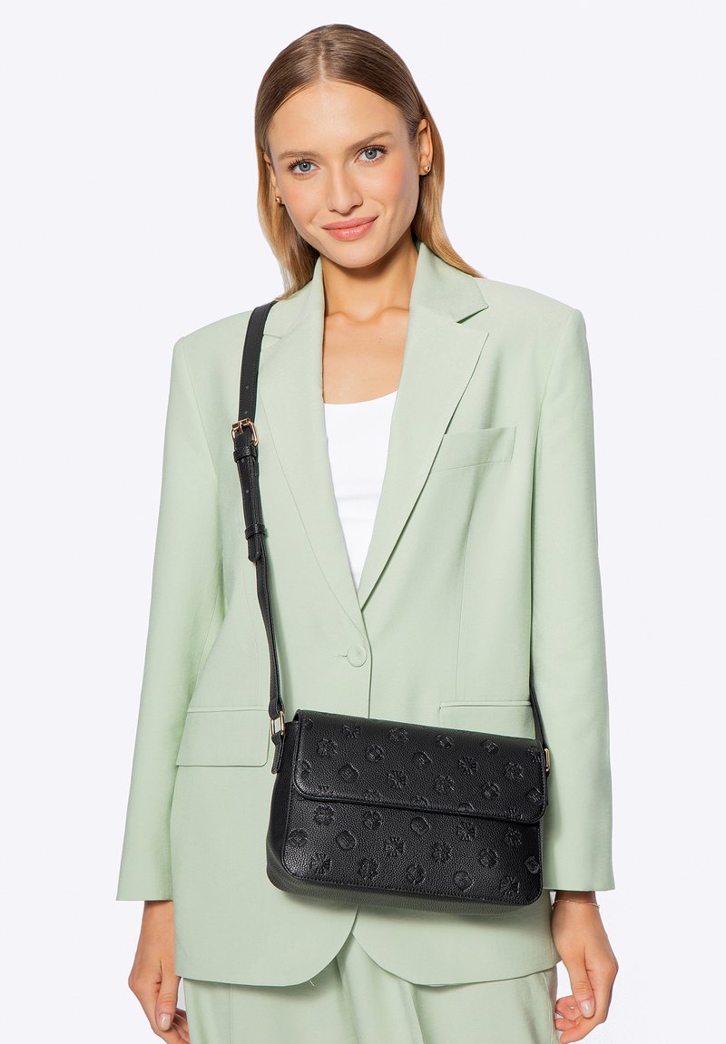 Schwarze, strukturierte Umhängetasche mit geprägten Mustern, verstellbarem Riemen und Klappverschluss. Getragen mit einem hellgrünen Blazer und weißem Oberteil.