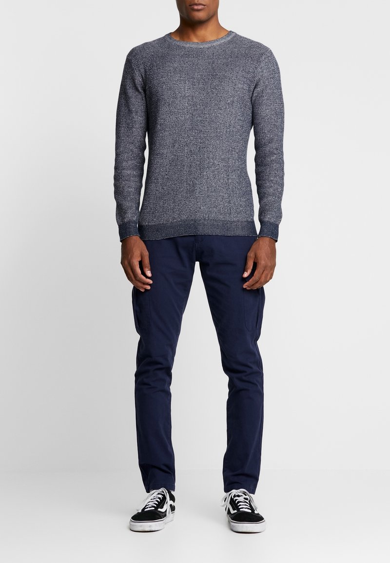 Marineblauwe gebreide trui met een gestructureerde afwerking, gecombineerd met marineblauwe cargobroek en zwart-witte sneakers met veters en een lage zool.