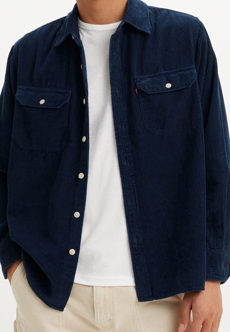 Homme portant une chemise en velours côtelé bleu marine boutonnée sur un t-shirt blanc et un pantalon beige, les mains dans les poches, torse visible.