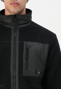 Bărbat îmbrăcat cu o jachetă de fleece neagră, cu guler înalt și un buzunar pe piept cu fermoar, având o etichetă mică neagră.