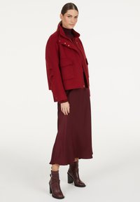 Frau in einem burgunderfarbenen Rollkragenpullover, einem passenden Midirock, einer Wolljacke mit großen Taschen und dunkellbraunen, hochhackigen Stiefeletten.