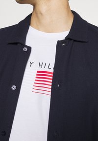 Chaqueta de camisa de punto azul marino con dos botones negros y cuello, llevada sobre una camiseta blanca con rayas rojas horizontales y texto gráfico.