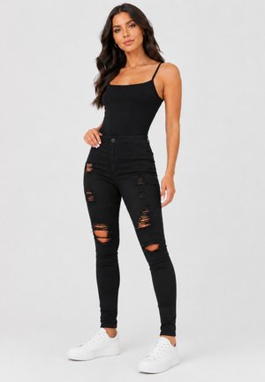 Kvinde med langt mørkt hår iført en sort spaghetti-strop top, revnede sorte skinny jeans og hvide sneakers, der står mod en grå baggrund.