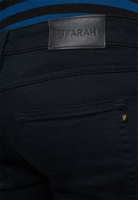 Pantalones vaqueros negros con un parche de cuero etiquetado "Farah." Presentan costuras en contraste y un pequeño logo dorado en el bolsillo.