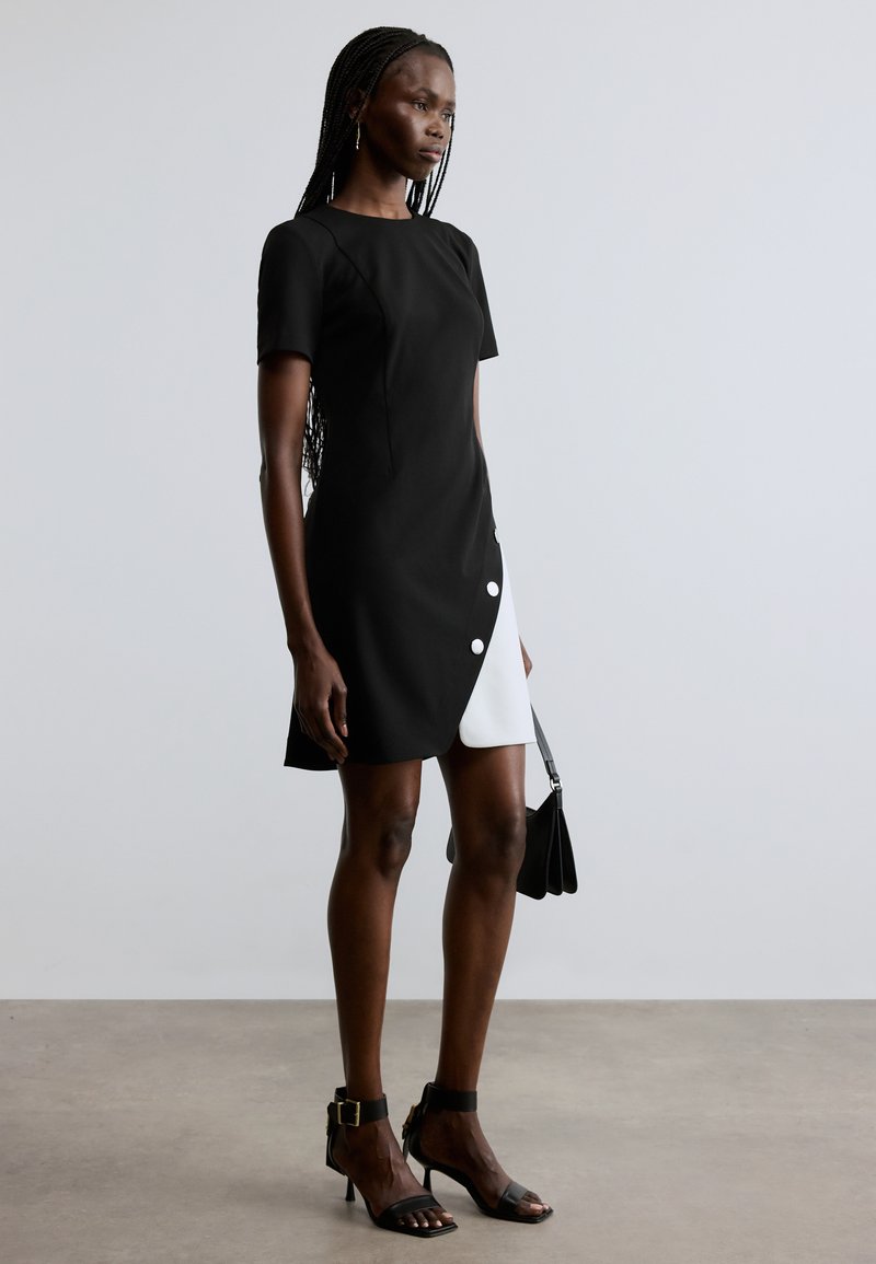 Vestido preto de manga curta com painel branco assimétrico, apresentando três grandes botões brancos na lateral. Usado com sandálias pretas de salto alto.