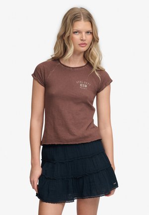 Jeune femme aux cheveux blonds portant un t-shirt marron à manches courtes et une jupe noire à volants, debout devant un fond blanc.