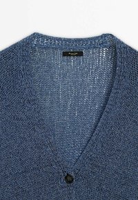 Blå stickad kofta med V-ringning, lös vävstruktur och en svart knapp framtill. Etikett synlig inuti.