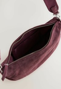 Bordeaux ruskind taske med lynlås på toppen, stofrem og en indre lomme. Den bløde tekstur og det minimalistiske design fremhæver funktionaliteten.