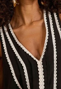 Svart ärmlös topp i tyg med scallop-mönstrad vit spetskant längs halsringningen och skuldersömmarna, med vertikala veckdetaljer.