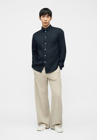 Άνδρας που φοράει σκουρομπλε πουκάμισο με κουμπιά, μπεζ παντελόνι με φαρδιά μπατζάκια και λευκά αθλητικά παπούτσια, στέκεται με τα χέρια στις τσέπες σε απλό φόντο.