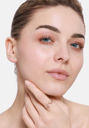 Bague en or rose en forme de vague sur une main, texture lisse, courbes douces, portée sur un doigt près du menton, avec un maquillage rose subtil et une peau claire.