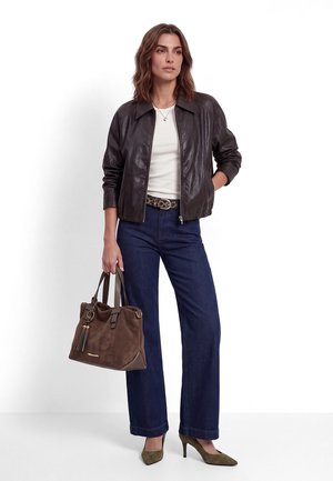 Donna che indossa giacca in pelle marrone scuro, camicia bianca, jeans blu, tacchi verdi, che tiene in mano una borsa in camoscio marrone con dettaglio a nappine.