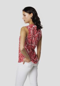 Top rojo sin mangas con un estampado de hojas blancas, confeccionado en tela ligera, con dobladillo curvado y ajuste relajado, combinado con pantalones blancos.