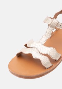 Καφέ επίπεδη σαγιονάρα με λαμπερές, λευκές λουρίδες με κυματιστό σχέδιο φίδι και κούμπωμα σε λευκό φόντο.