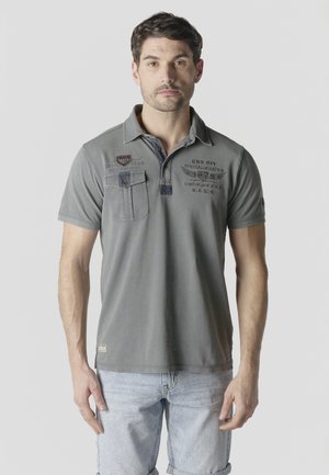 Hombre con polo gris con diseño gráfico y pantalones cortos de mezclilla azul claro remangados, de pie contra un fondo liso.