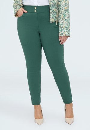 Donna che indossa pantaloni verdi a vita alta e aderenti con due bottoni, una giacca con motivo, una camicia bianca e tacchi beige appuntiti.